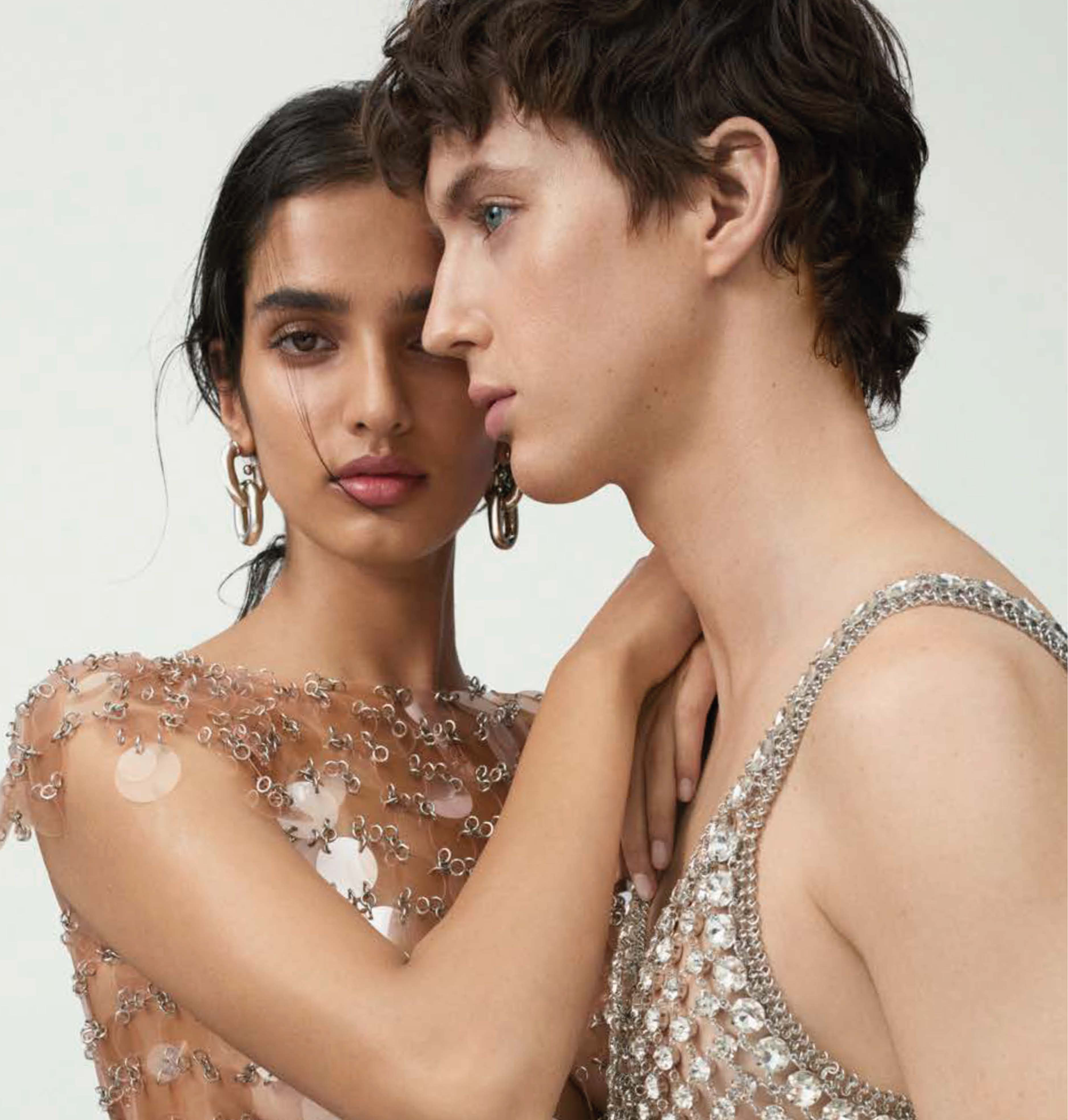 Fashion portrait of a fair-skinned adult male and a tan-skinned adult female, embracing in chainmail-style dresses against a white backdrop.