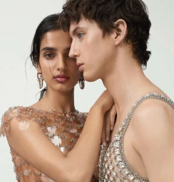 Close-up portrait of two models embracing against a seamless white background, wearing ornate embellished garments and drop earrings.