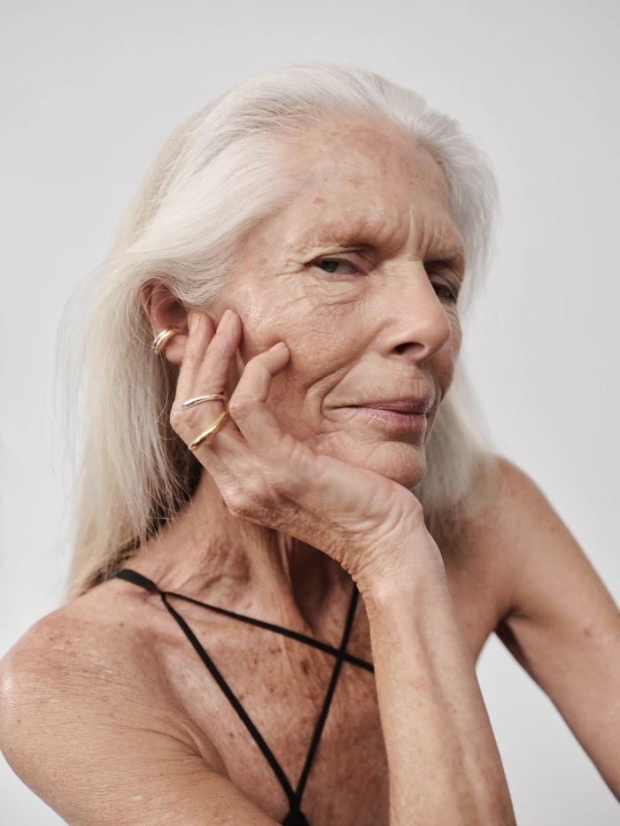 Elderly woman with long white hair wearing gold rings and an ear cuff with thin black straps against a seamless background