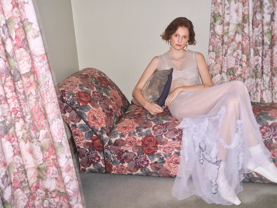Woman in sheer white dress seated on a floral chaise lounge framed by matching floral curtains