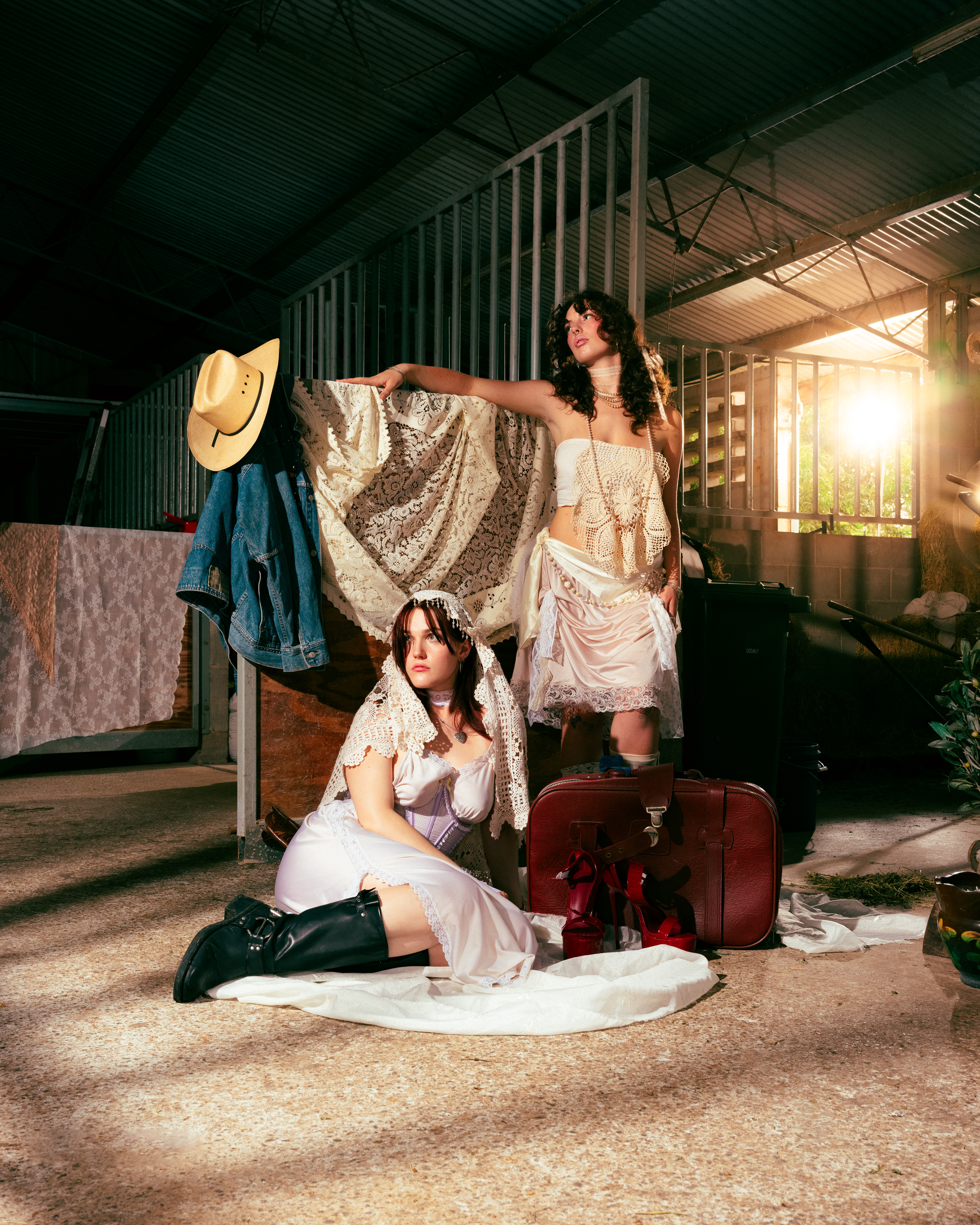 Two women in vintage-inspired attire pose in a rustic barn, one seated with black boots, the other standing with a cream-colored shawl.