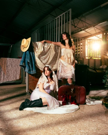 Two women in vintage-inspired attire pose in a rustic barn, one seated with black boots, the other standing with a cream-colored shawl.