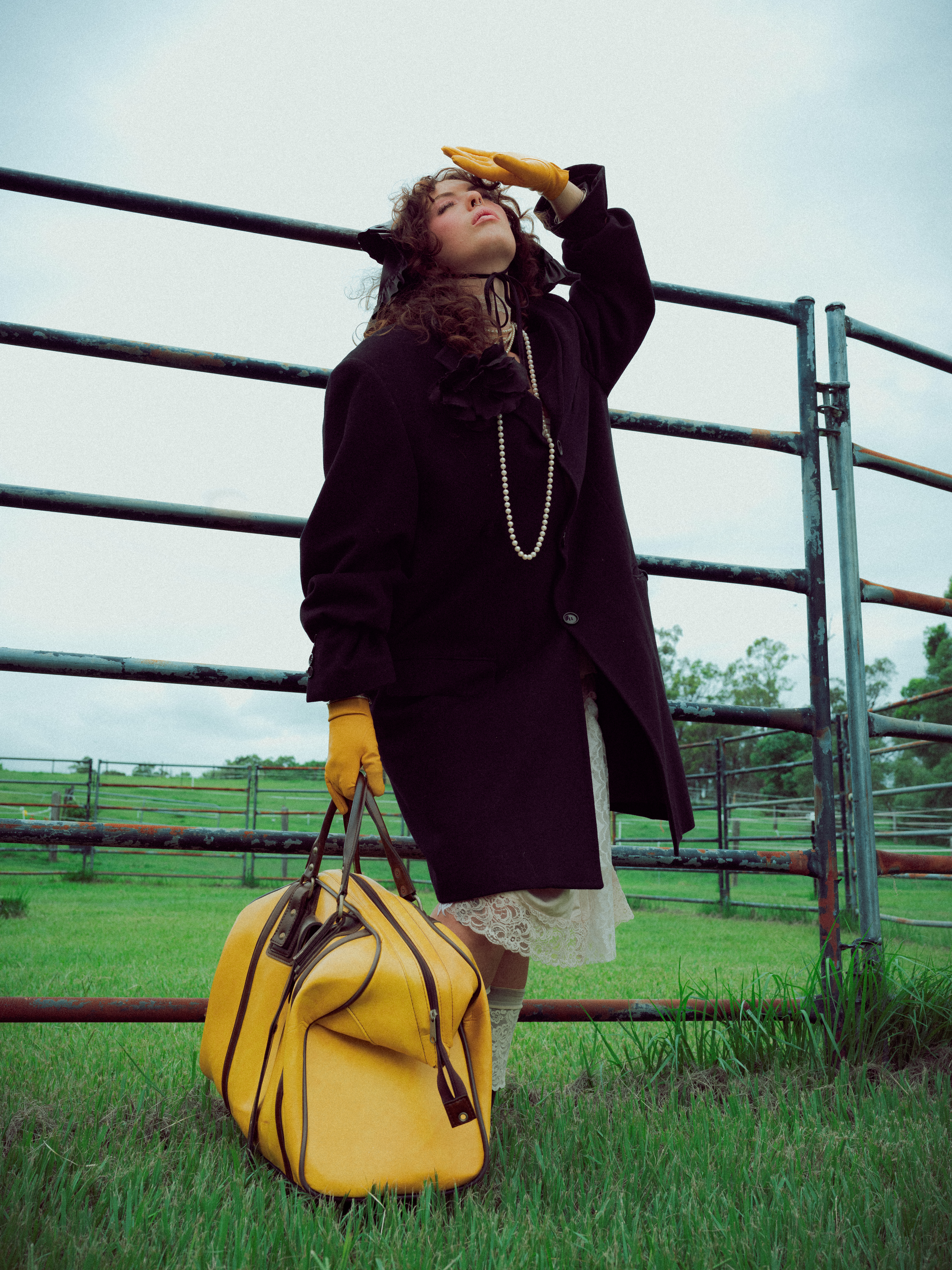 Woman in coat stands near metal fence with yellow duffel bag in a grassy field, wearing yellow gloves and pearls with overcast sky.