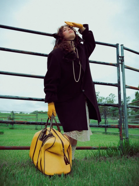 Woman in coat stands near metal fence with yellow duffel bag in a grassy field, wearing yellow gloves and pearls with overcast sky.