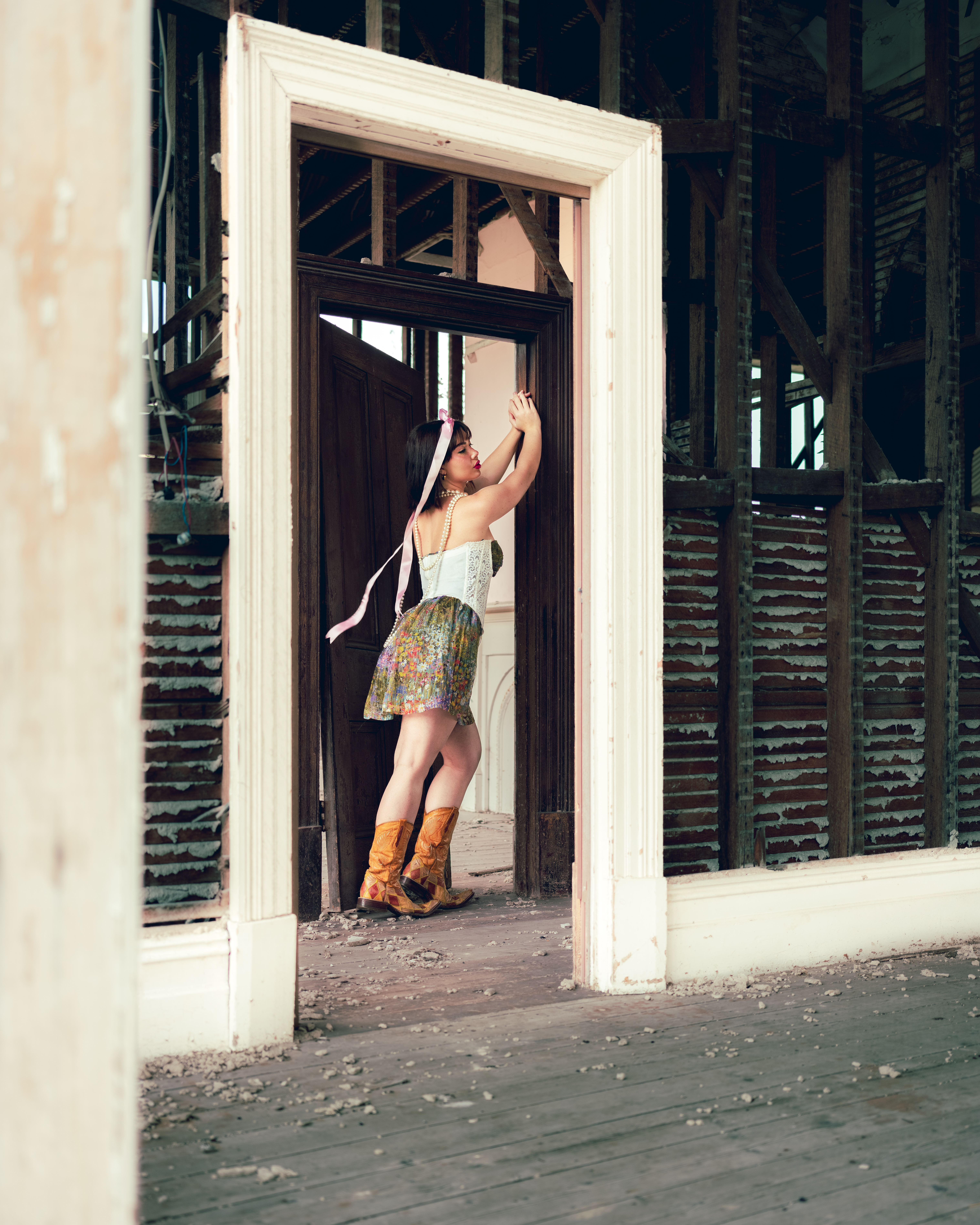Young woman poses in a floral skirt, cowboy boots, and corset within a dilapidated home interior, standing within an empty doorway.