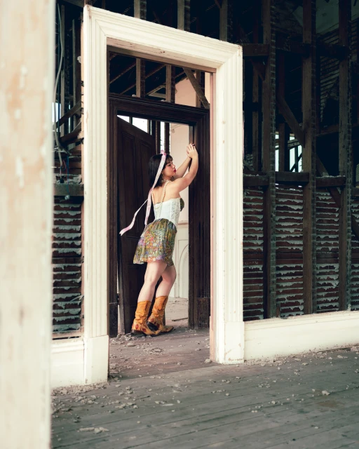 Young woman poses in a floral skirt, cowboy boots, and corset within a dilapidated home interior, standing within an empty doorway.
