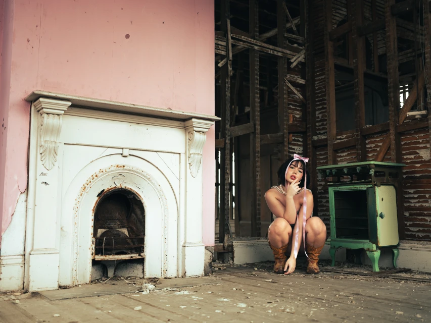 Woman in vintage boots and pearl necklace poses next to antique stove in abandoned room, pink walls, natural light, and distressed flooring.
