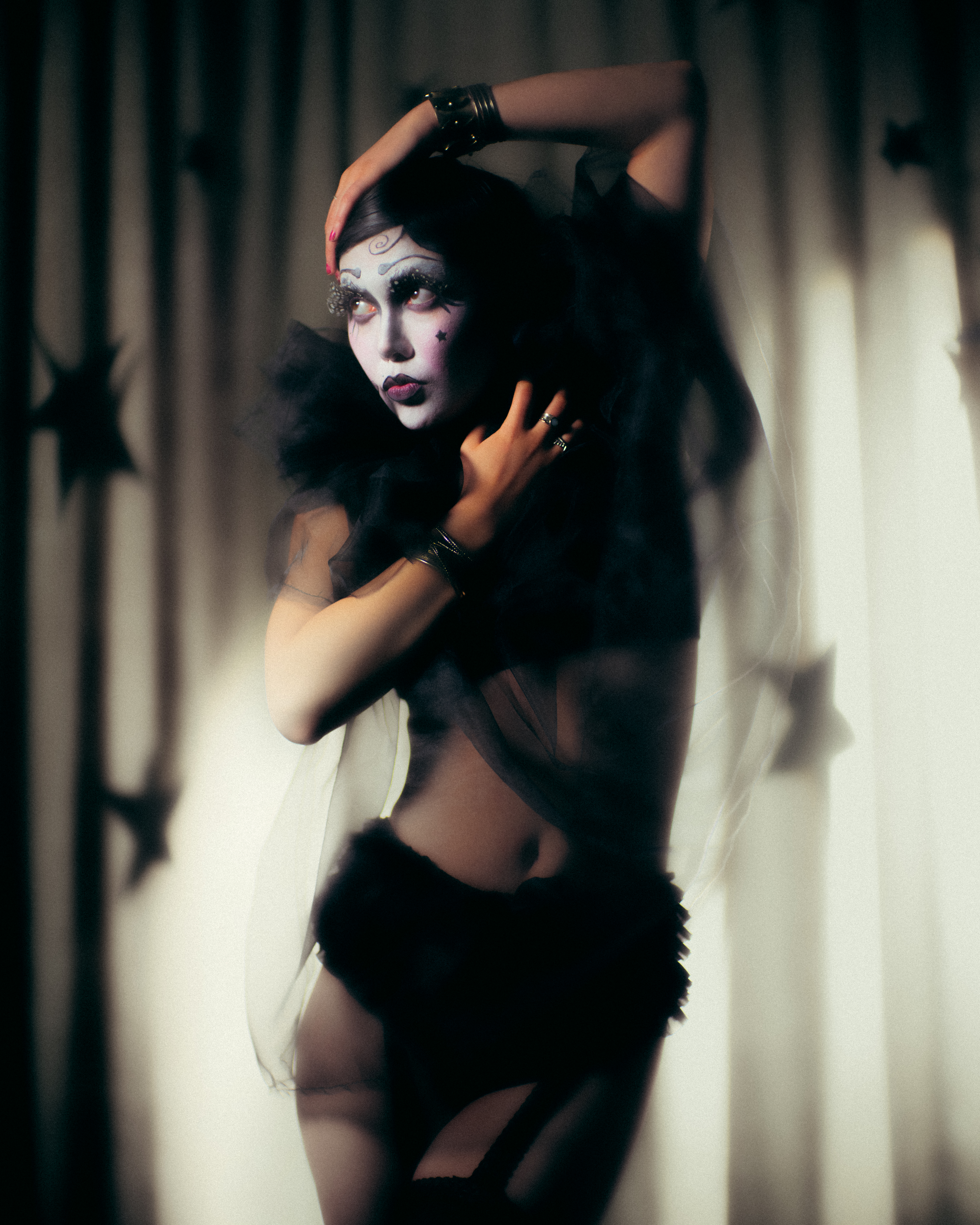 Woman in theatrical makeup poses in sheer lingerie against a star-studded curtain, creating a dark, boudoir-inspired fashion image.