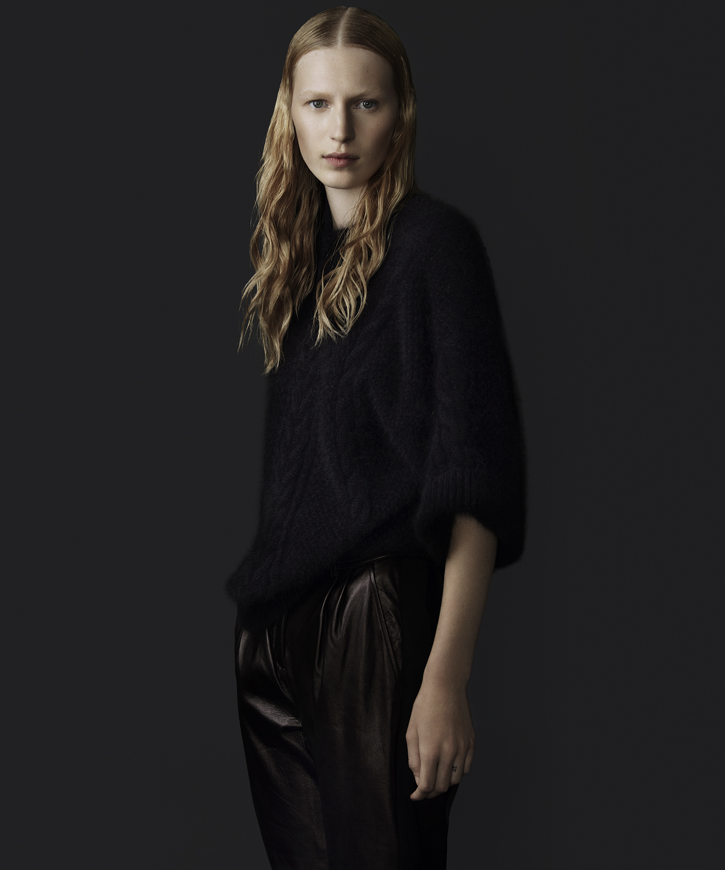 A blonde adult woman poses in a black fuzzy sweater and dark shiny pants against a solid dark background in a studio shot.