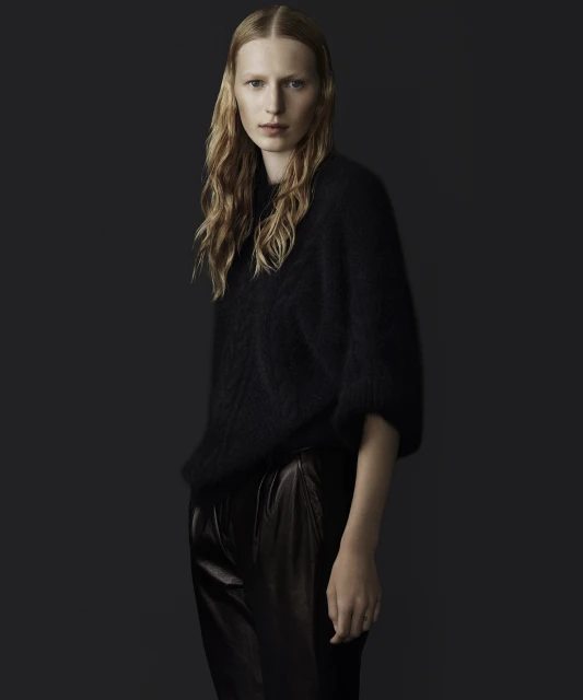 A blonde adult woman poses in a black fuzzy sweater and dark shiny pants against a solid dark background in a studio shot.