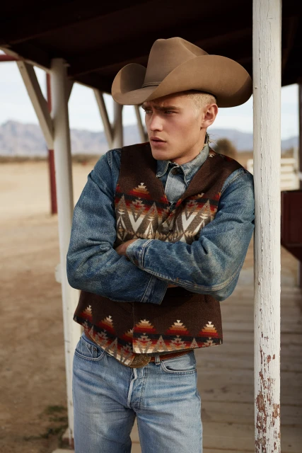 Man in cowboy hat and vest stands outside leaning against a post with arms crossed in a rural desert landscape, looking away.