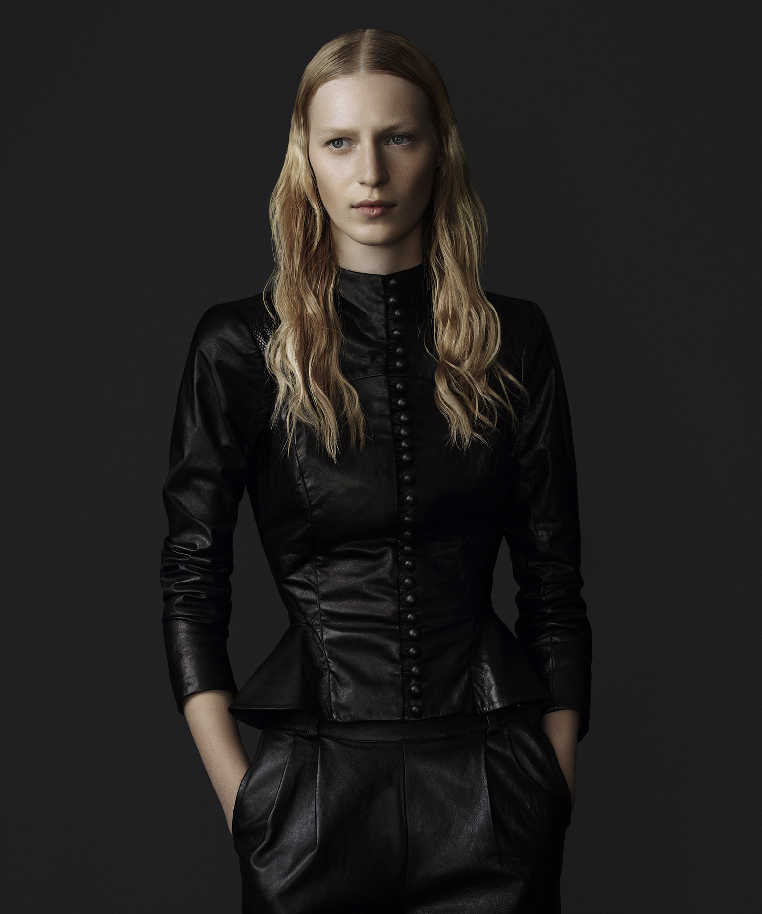 Blonde woman in a long-sleeved black leather top with a high neckline and peplum, and black leather pants posing against a grey background.