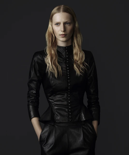 Blonde woman in a long-sleeved black leather top with a high neckline and peplum, and black leather pants posing against a grey background.