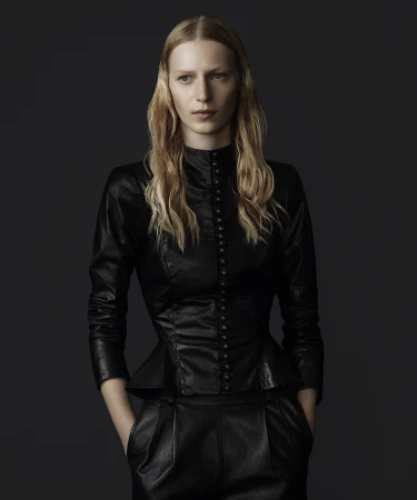 Blonde woman in a long-sleeved black leather top with a high neckline and peplum, and black leather pants posing against a grey background.