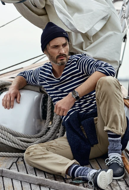 Adult male model posing on a sailboat wearing a navy blue beanie, striped shirt, khaki pants, and navy boat shoes with grey cloudy sky in background.