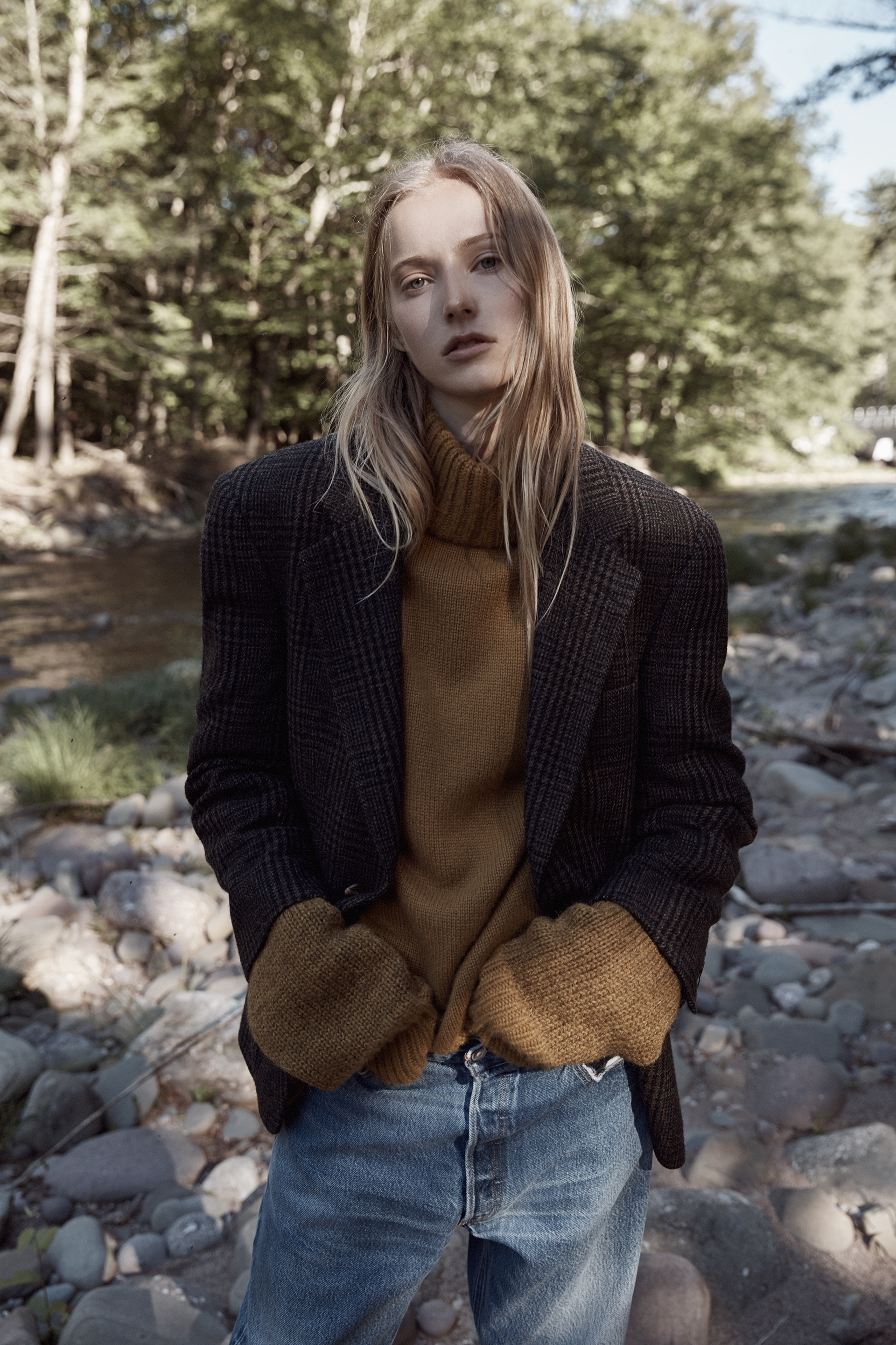 A blonde woman stands in an outdoor setting, wearing a blazer, a turtleneck sweater, and blue jeans, with a serene expression.