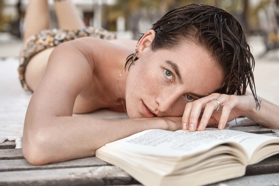 Adult woman lying down reading a book outside on a wooden surface with a blurred background of beach trees.