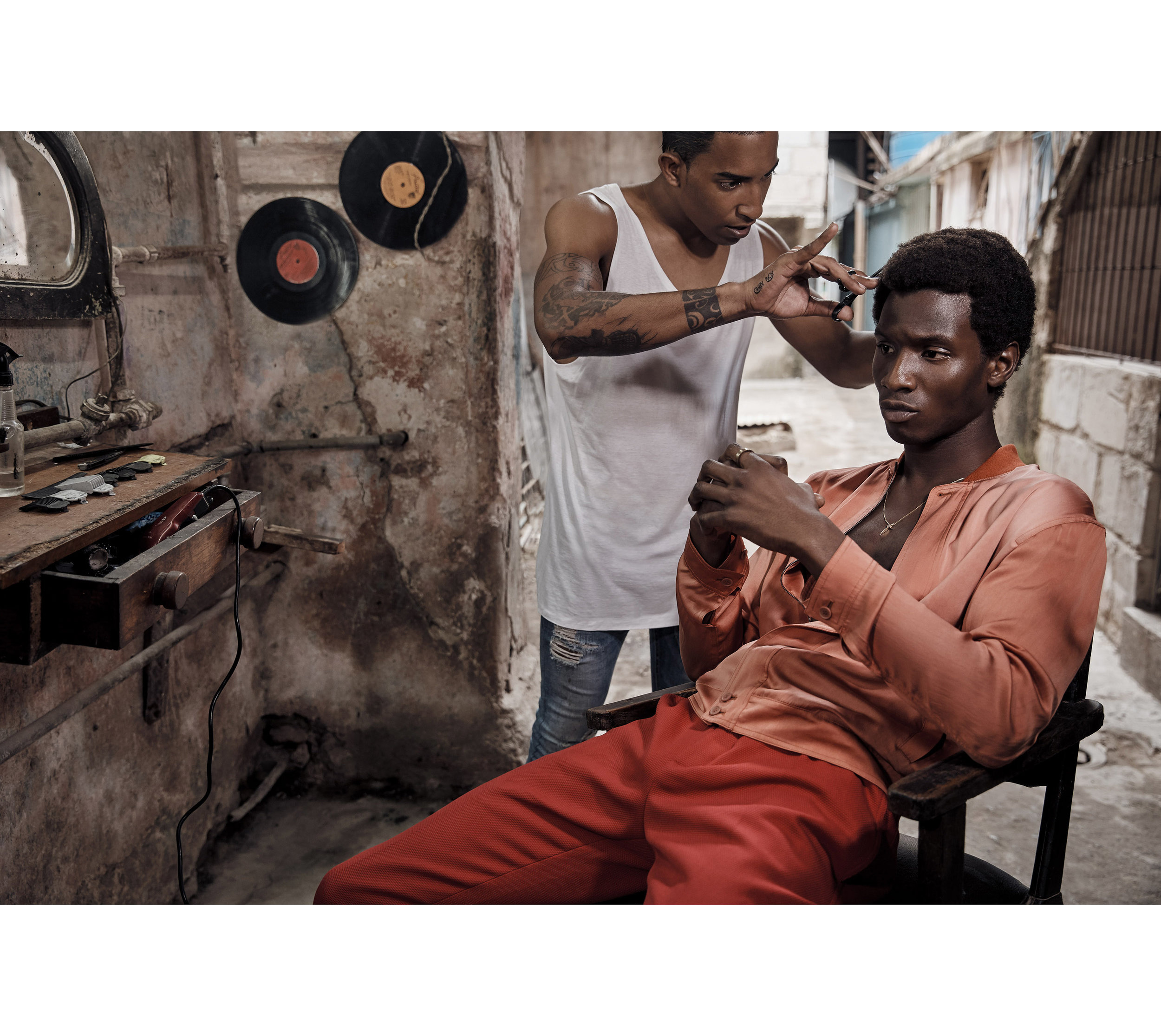Young man getting a haircut from a barber in an indoor barbershop. The client is sitting in a chair, wearing an orange shirt and pants.