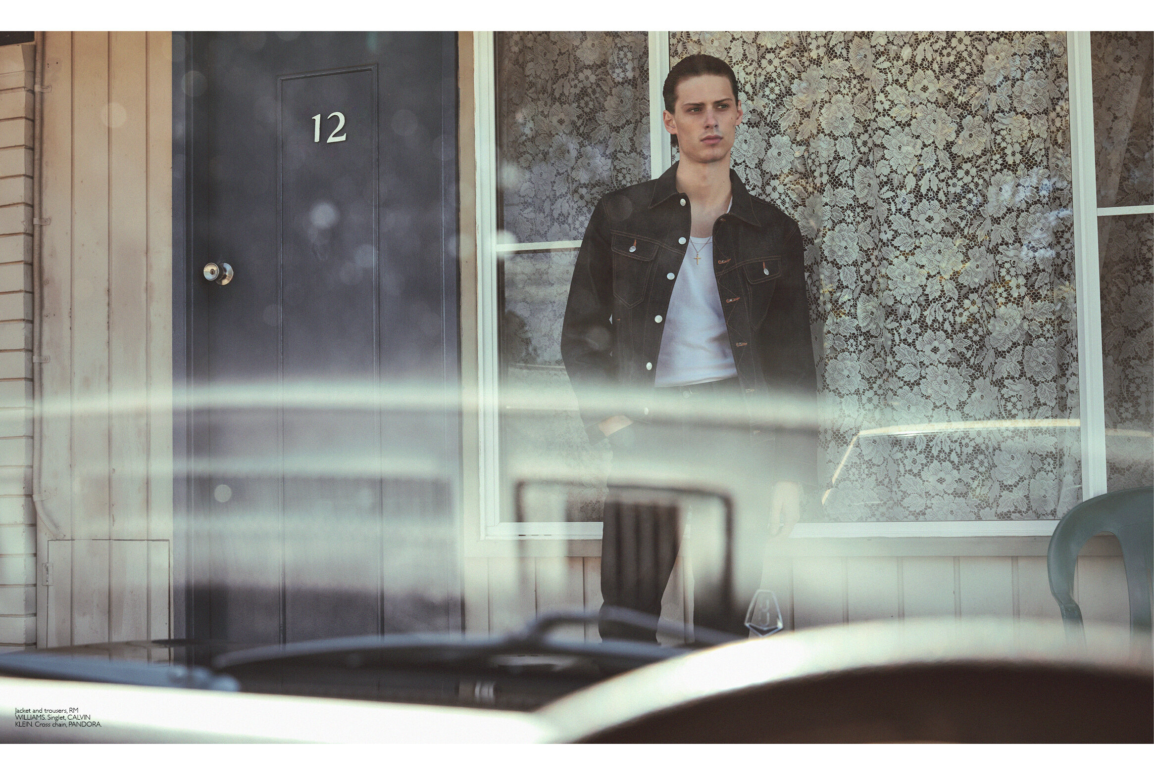A stylish young adult stands in a doorway with a dark denim jacket and a white t-shirt, reflected in a car windshield.