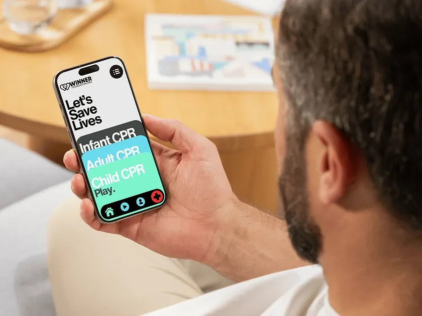 Adult man holds a smartphone displaying a CPR training app with options for infant, adult, and child CPR, set against a blurred indoor background.