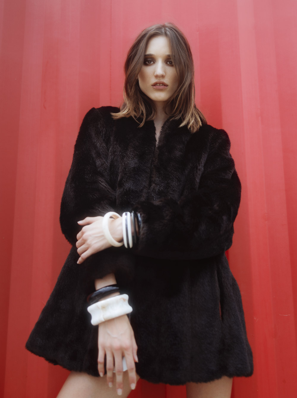 A woman in a black fur coat poses against a red wall, showcasing ivory and black wrist cuffs, with a serious expression and neutral hair.