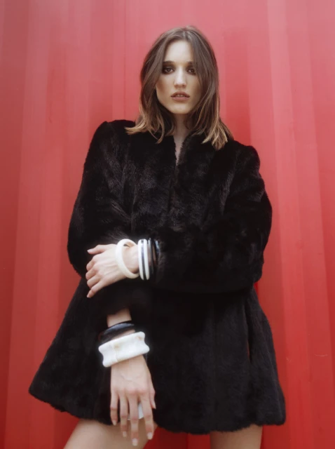 A woman in a black fur coat poses against a red wall, showcasing ivory and black wrist cuffs, with a serious expression and neutral hair.