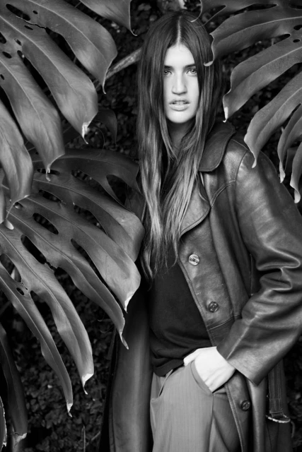 A woman with long brown hair stands in front of large monstera leaves, wearing a dark leather trench coat and trousers in a studio portrait.