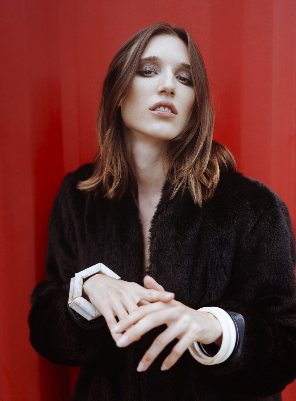 Stylish woman with shoulder-length brown hair poses in a black fur coat against a vibrant red background, wearing unique white bracelets.