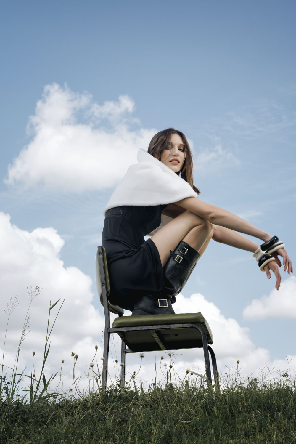 A woman in a dark dress and boots poses on a chair in a grassy field under a blue sky with white clouds, fashion editorial.