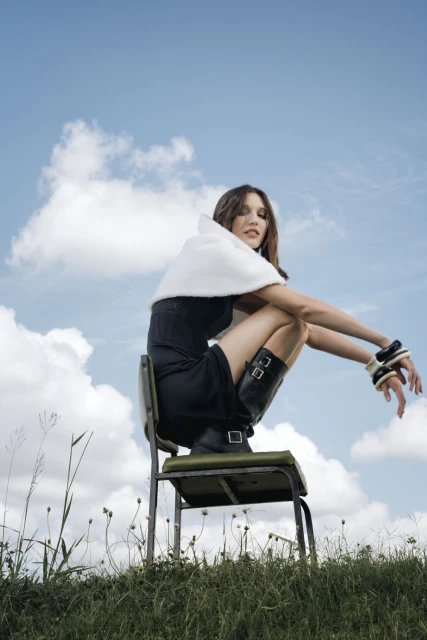 A woman in a dark dress and boots poses on a chair in a grassy field under a blue sky with white clouds, fashion editorial.