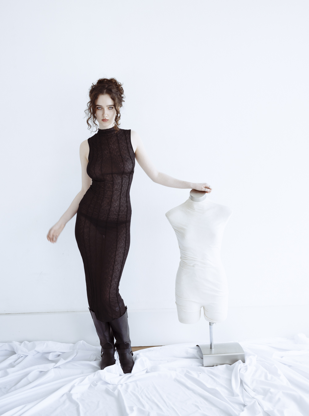 Woman in a sheer black sleeveless dress and dark boots standing next to a white mannequin torso.