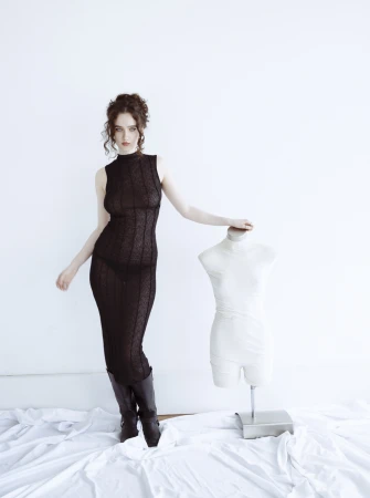 Woman in a sheer black sleeveless dress and dark boots standing next to a white mannequin torso.