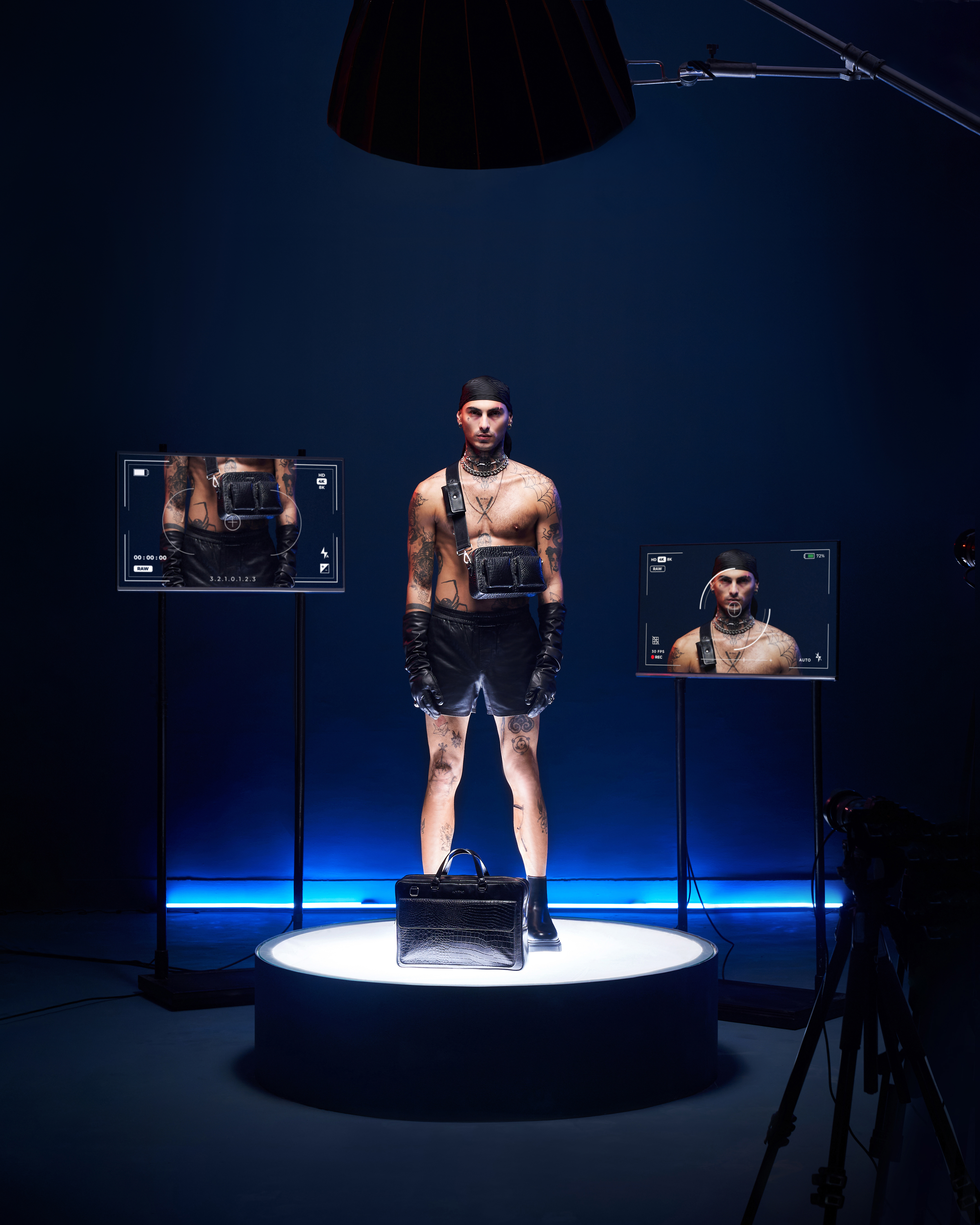 Male fashion model in leather shorts stands on platform in studio, displayed on screens in photo studio with camera visible.