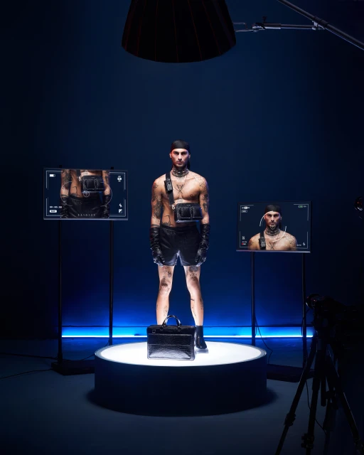 Male fashion model in leather shorts stands on platform in studio, displayed on screens in photo studio with camera visible.