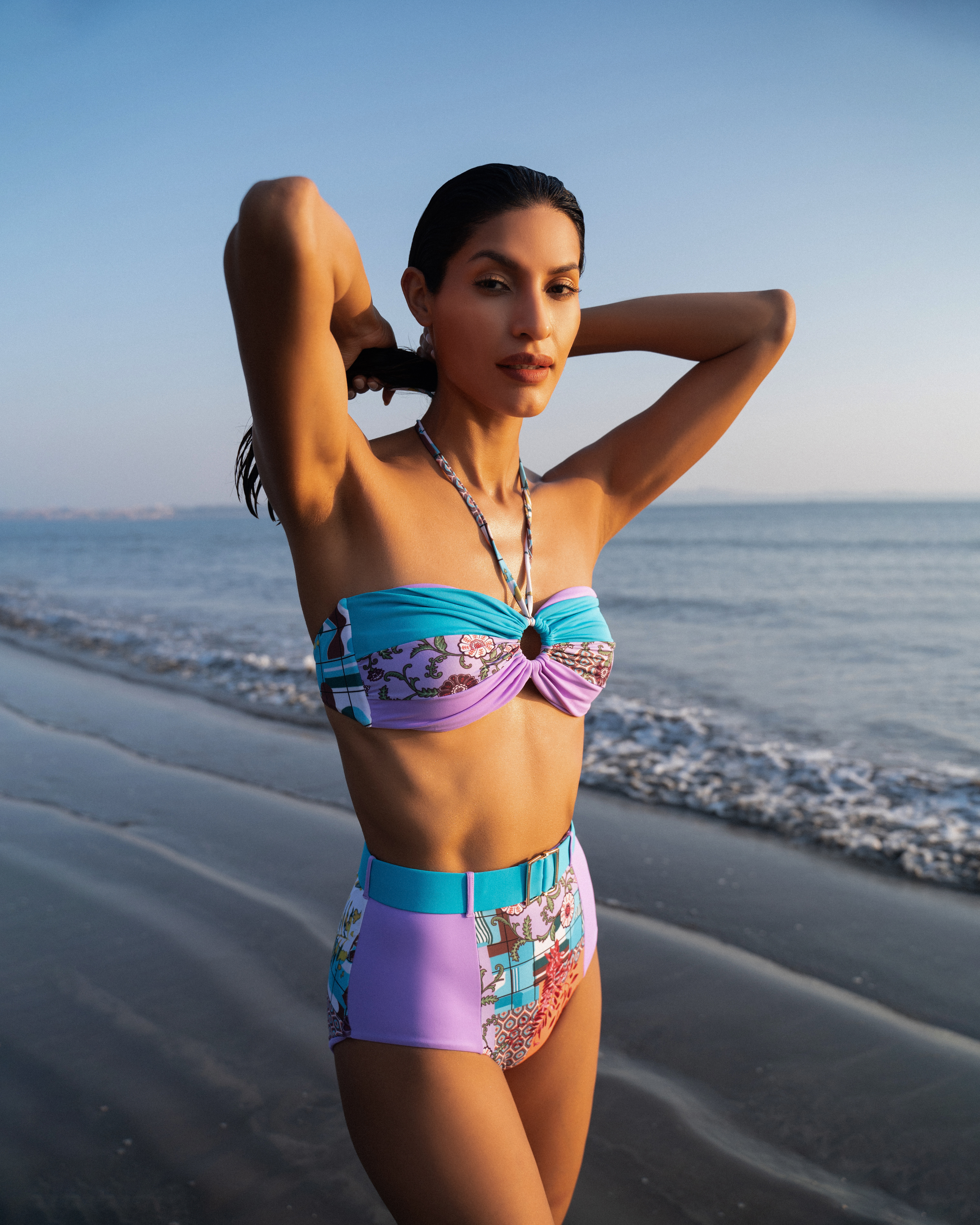 Woman poses on beach in stylish printed bikini with hands in hair, featuring sandy shoreline and ocean backdrop under bright daylight.