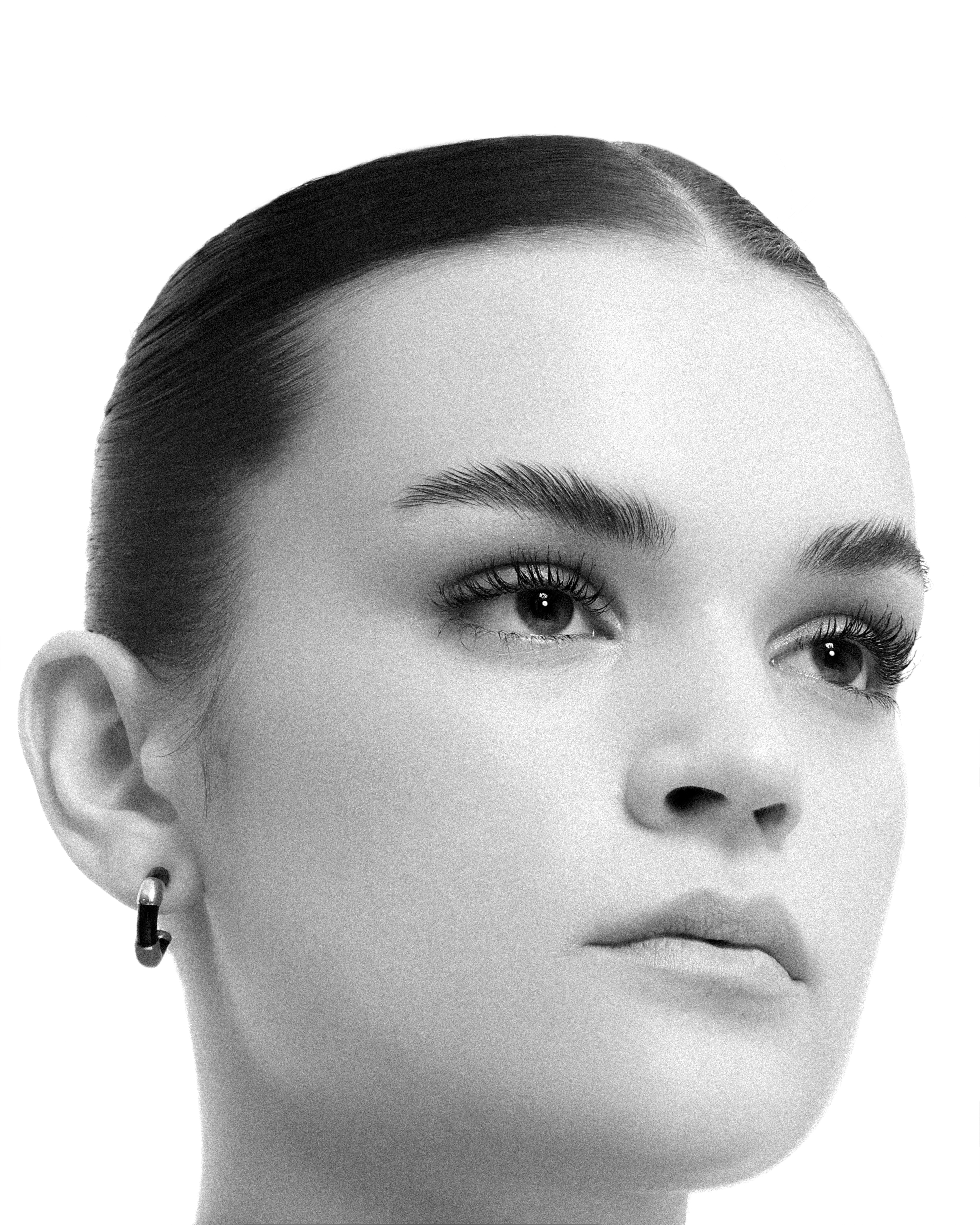 Close-up portrait of a fair-skinned adult woman with dark hair slicked back, featuring simple hoop earrings against a white background.