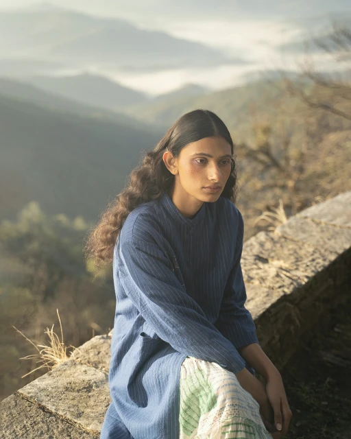 Woman with long dark hair poses in blue dress on rocky outcrop, overlooking mist-shrouded mountains during daytime in fashion-style shot.