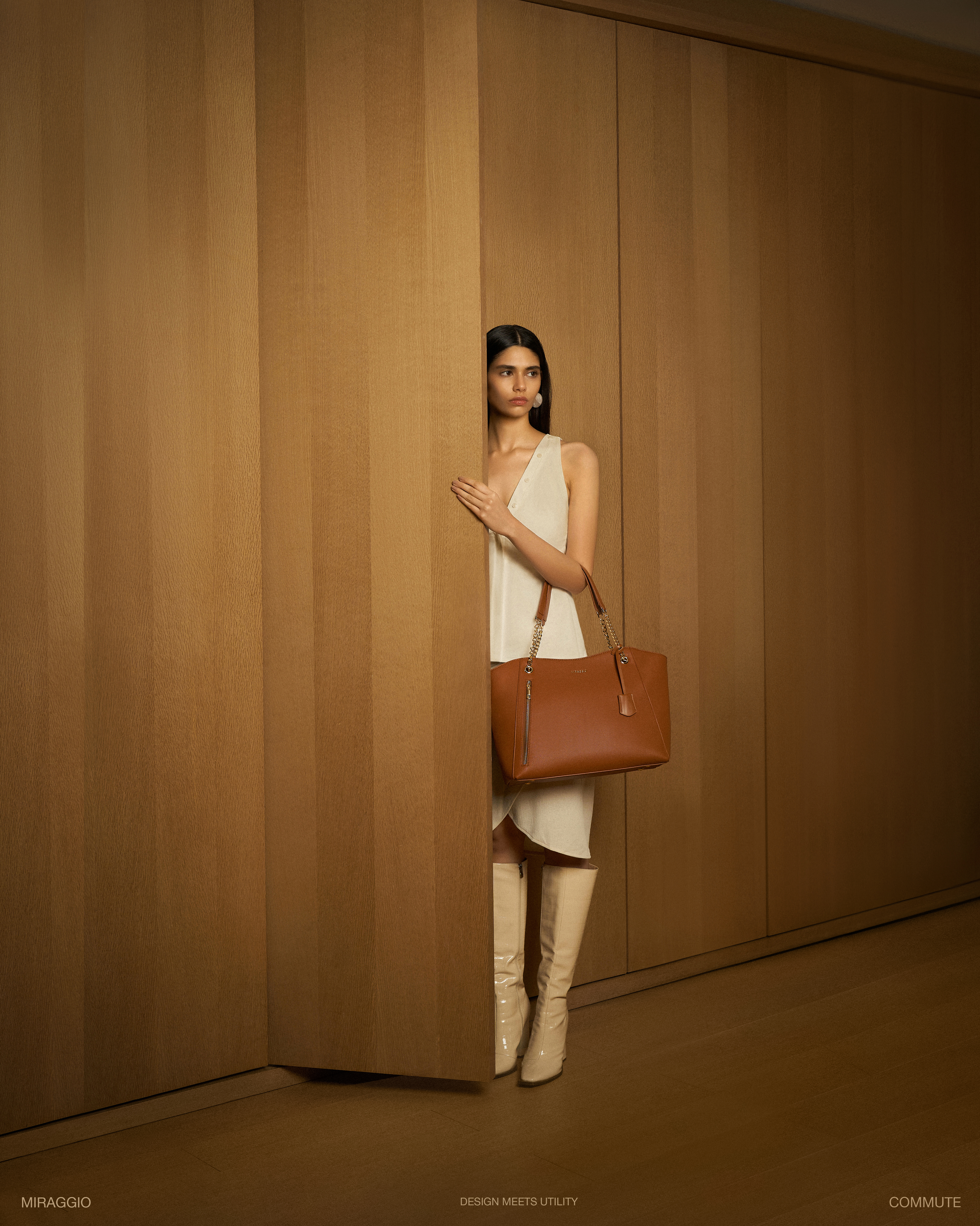 Fashion shot of a woman wearing a white dress, cream-colored boots, and carrying a brown leather tote bag, posing in front of a wood wall.