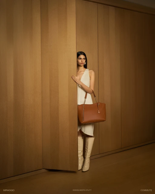 Fashion shot of a woman wearing a white dress, cream-colored boots, and carrying a brown leather tote bag, posing in front of a wood wall.