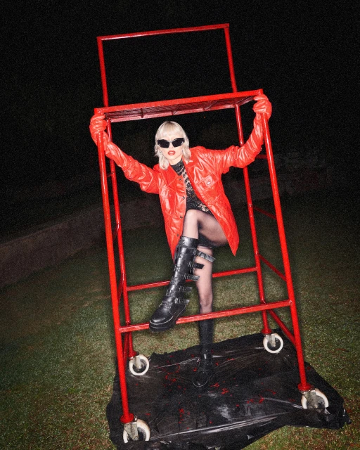 Woman in a red jacket and black boots poses on a red rolling scaffolding platform in an outdoor nighttime setting with dark, grassy ground.