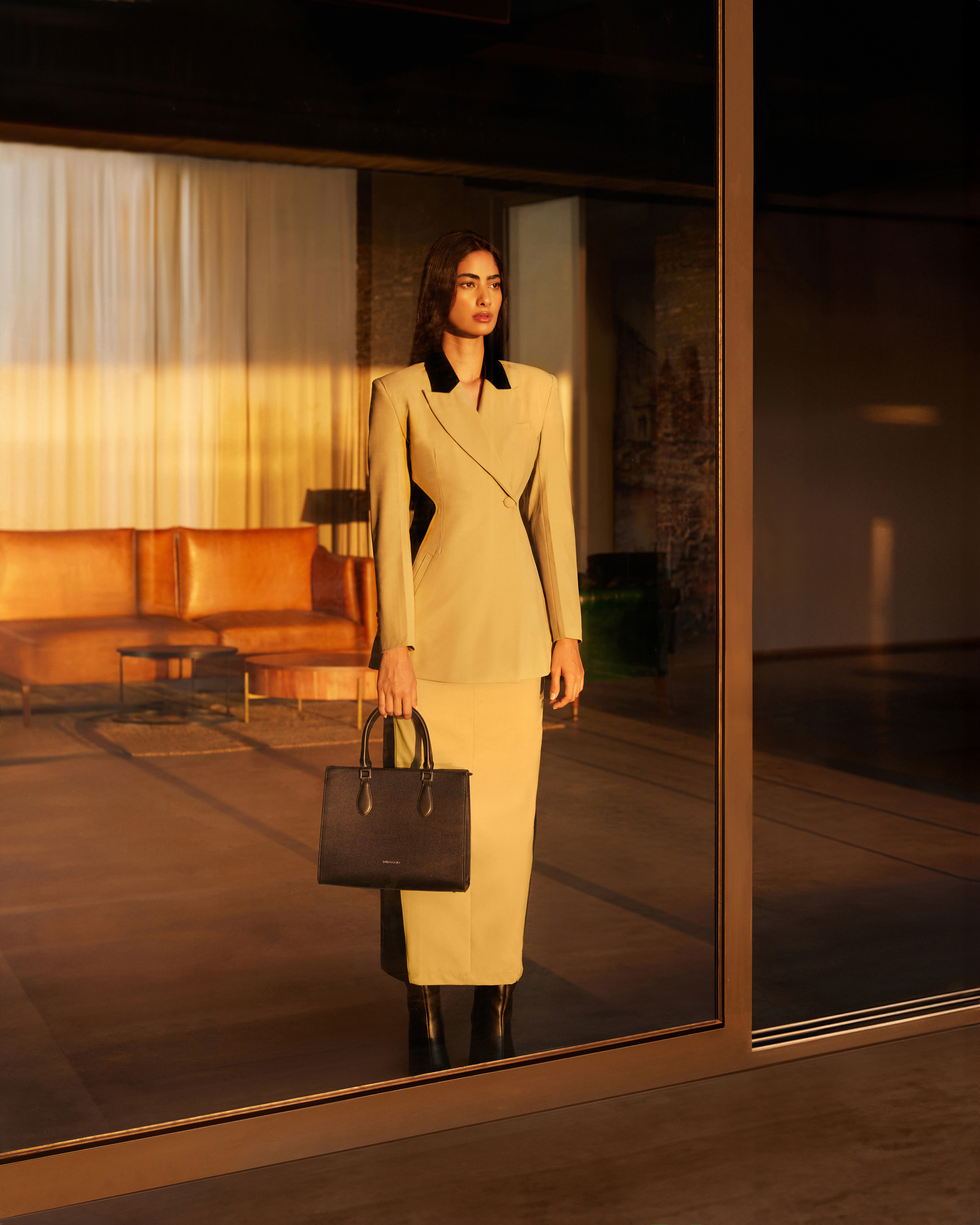 A woman in a tan skirt suit and black handbag is framed by a reflective glass doorway inside an interior space with tan furnishings.
