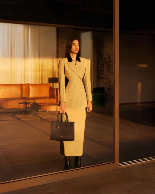 A woman in a tan skirt suit and black handbag is framed by a reflective glass doorway inside an interior space with tan furnishings.
