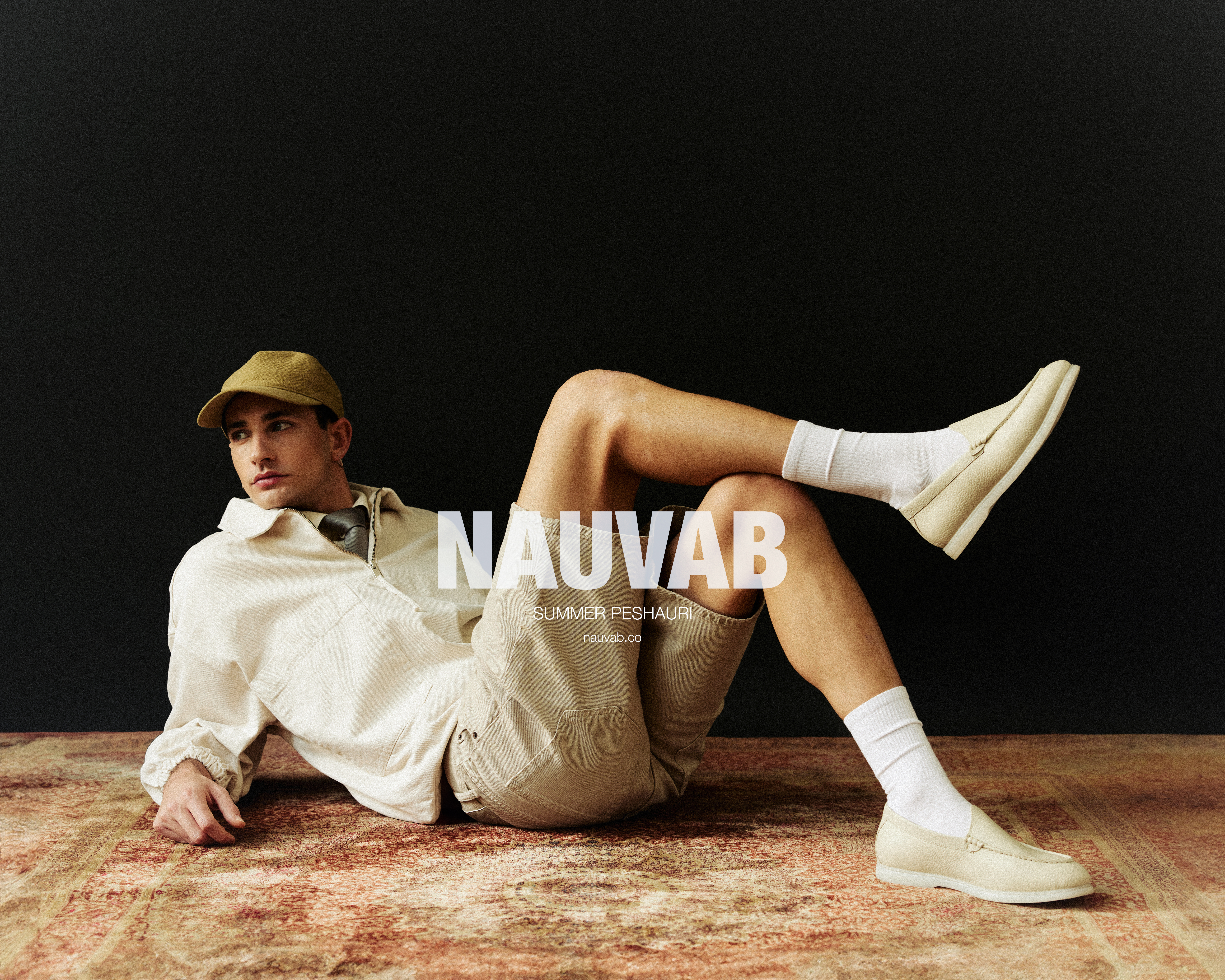 Fashion editorial shot of a young man in a beige outfit, cap, and off-white loafers, posing against a mottled stone wall and dark background.