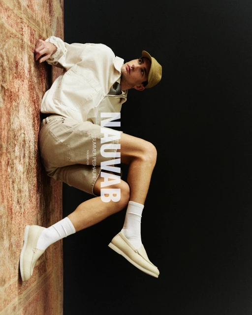 Fashion editorial shot of a young man in a beige outfit, cap, and off-white loafers, posing against a mottled stone wall and dark background.