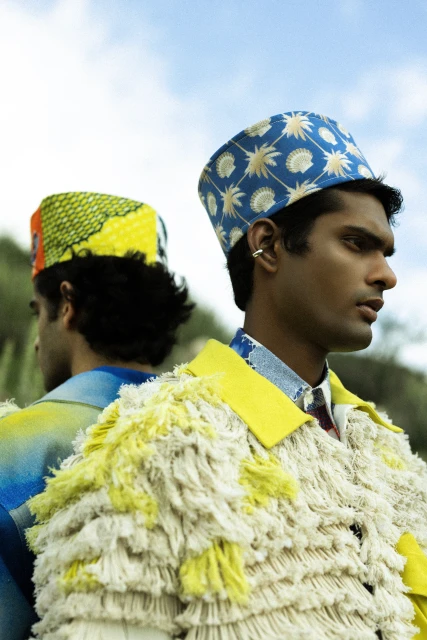Two stylish men in patterned hats and jackets stand back-to-back outdoors against a bright sky, showcasing unique fashion and design.