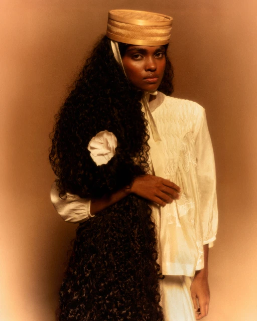 Portrait of a woman with dark curly hair wearing a white embroidered shirt and straw hat against a neutral backdrop in a studio.