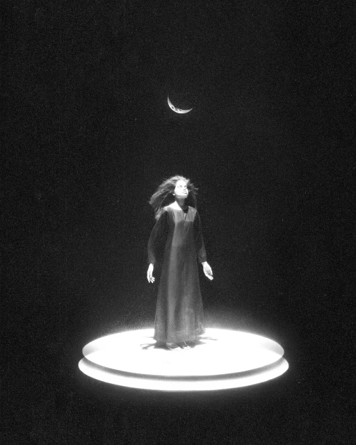Monochromatic photograph of a woman in a dark gown looking up at a crescent moon, standing on a lighted circular platform.