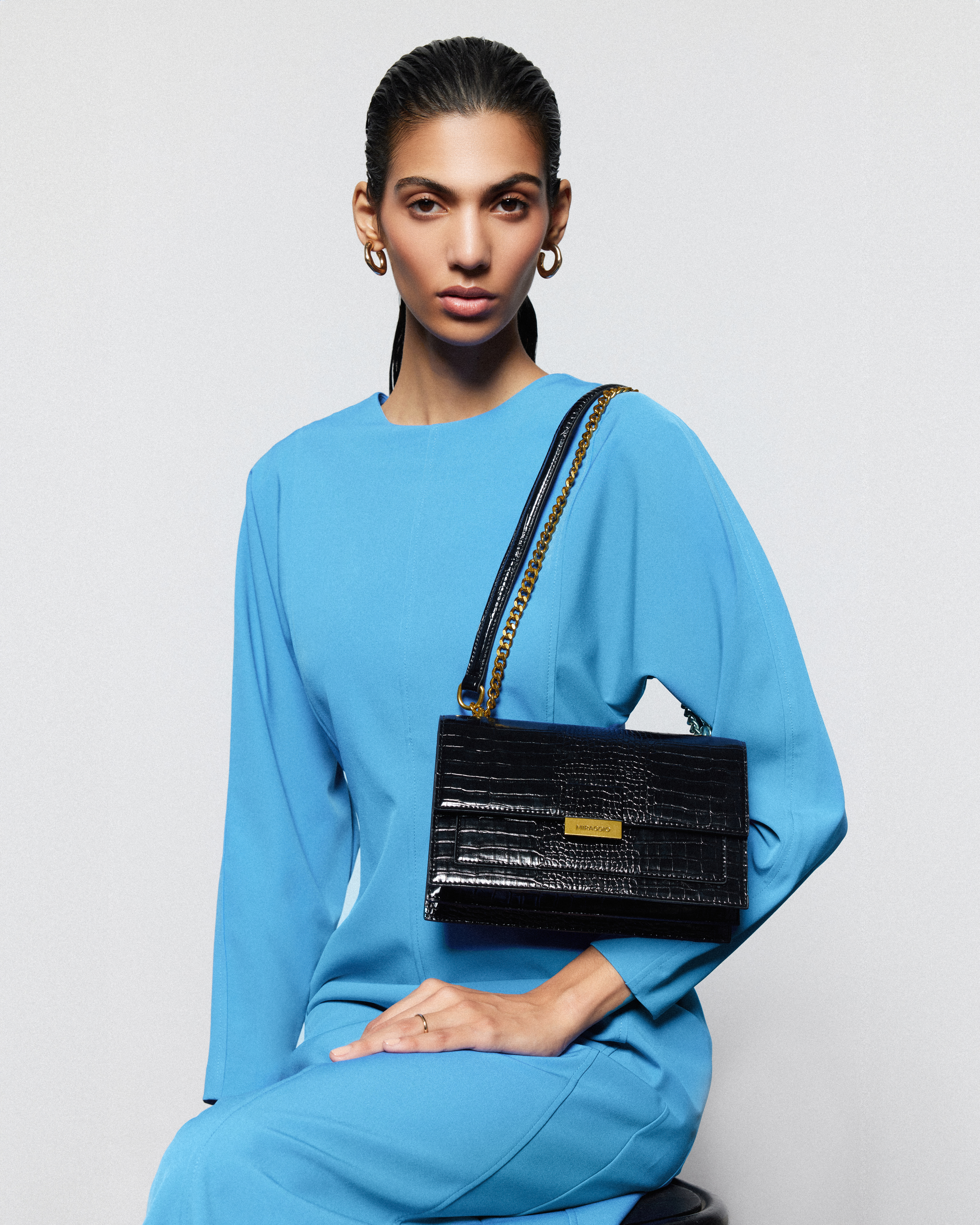 Adult female model seated in a photo studio wearing a turquoise dress and gold hoop earrings with a black crocodile handbag.