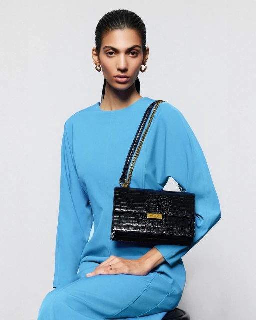 Adult female model seated in a photo studio wearing a turquoise dress and gold hoop earrings with a black crocodile handbag.