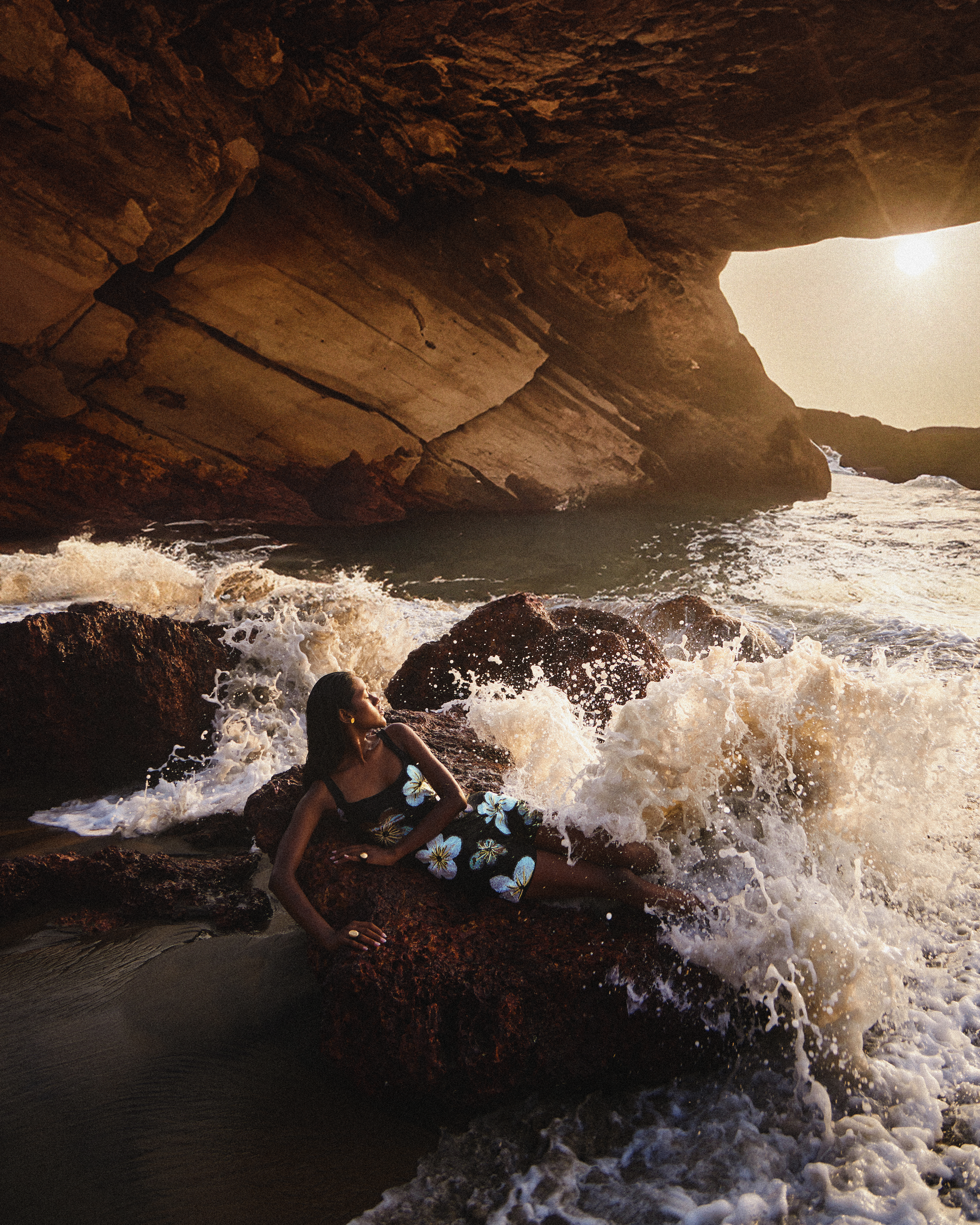 A woman in a floral dress lounges on rocks as waves crash around her, set against a cave entrance framing a sunset ocean view.